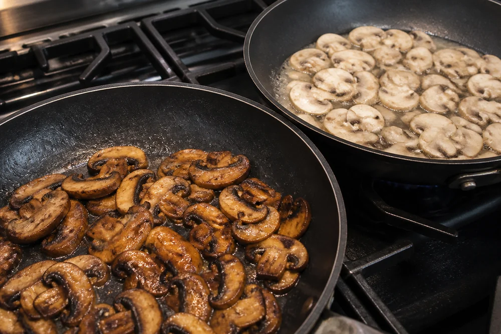 Sliced brown cremini mushrooms browning deeply in a hot skillet beside a second pan of paler white button slices releasing more liquid, both in one frame