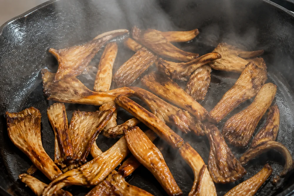Torn oyster mushroom strips crisped golden brown in a hot cast iron pan with dark curled edges and steam rising