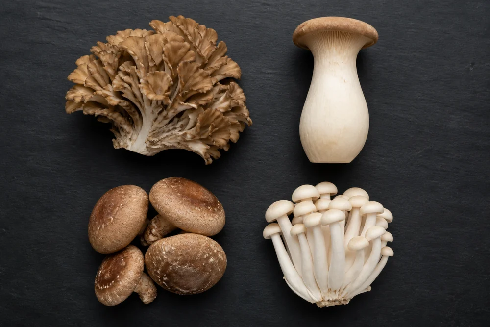 Four mushrooms in separate groups on a dark board, a frilled maitake cluster, a whole king oyster, several brown shiitake caps and a handful of white beech mushrooms