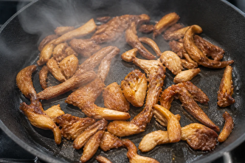 Torn pink oyster mushroom pieces frying in a hot skillet with the pink already faded to golden tan and brown at the crisped edges