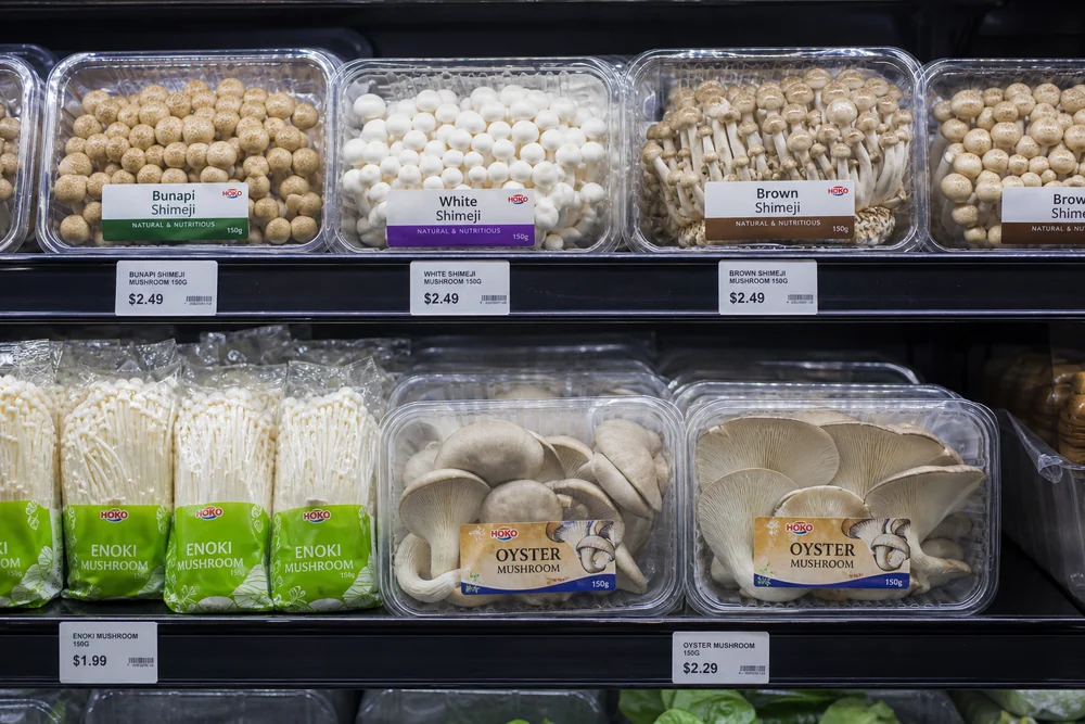 A chilled grocery shelf holding packs of shimeji, enoki and oyster mushrooms in clear punnets under cool lighting